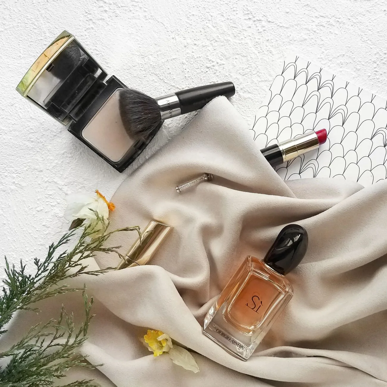 An arrangement of beauty products including a powder compact, perfume and lipstick on a soft cloth